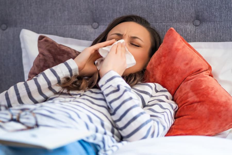 A woman laying in bed blowing her nose, emphasizing the need for hypoallergenic bed materials to reduce allergens.